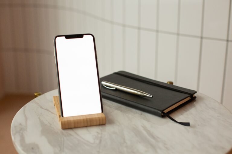 A smartphone with a blank screen on a wooden stand beside a closed notebook and pen.