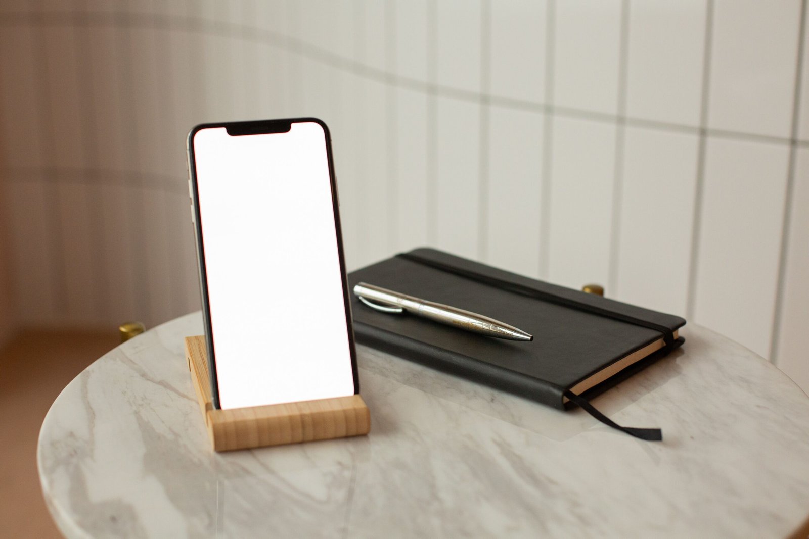 A smartphone with a blank screen on a wooden stand beside a closed notebook and pen.