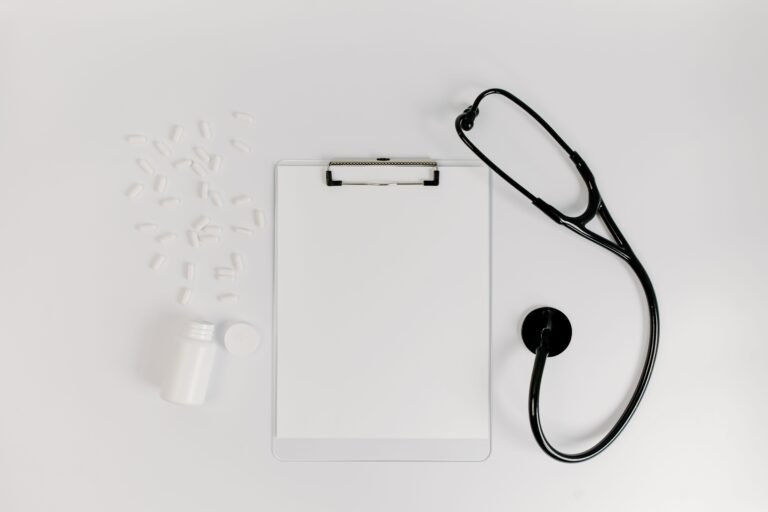 Clipboard, prescription bottle, pills, and stethoscope arranged on a white surface.