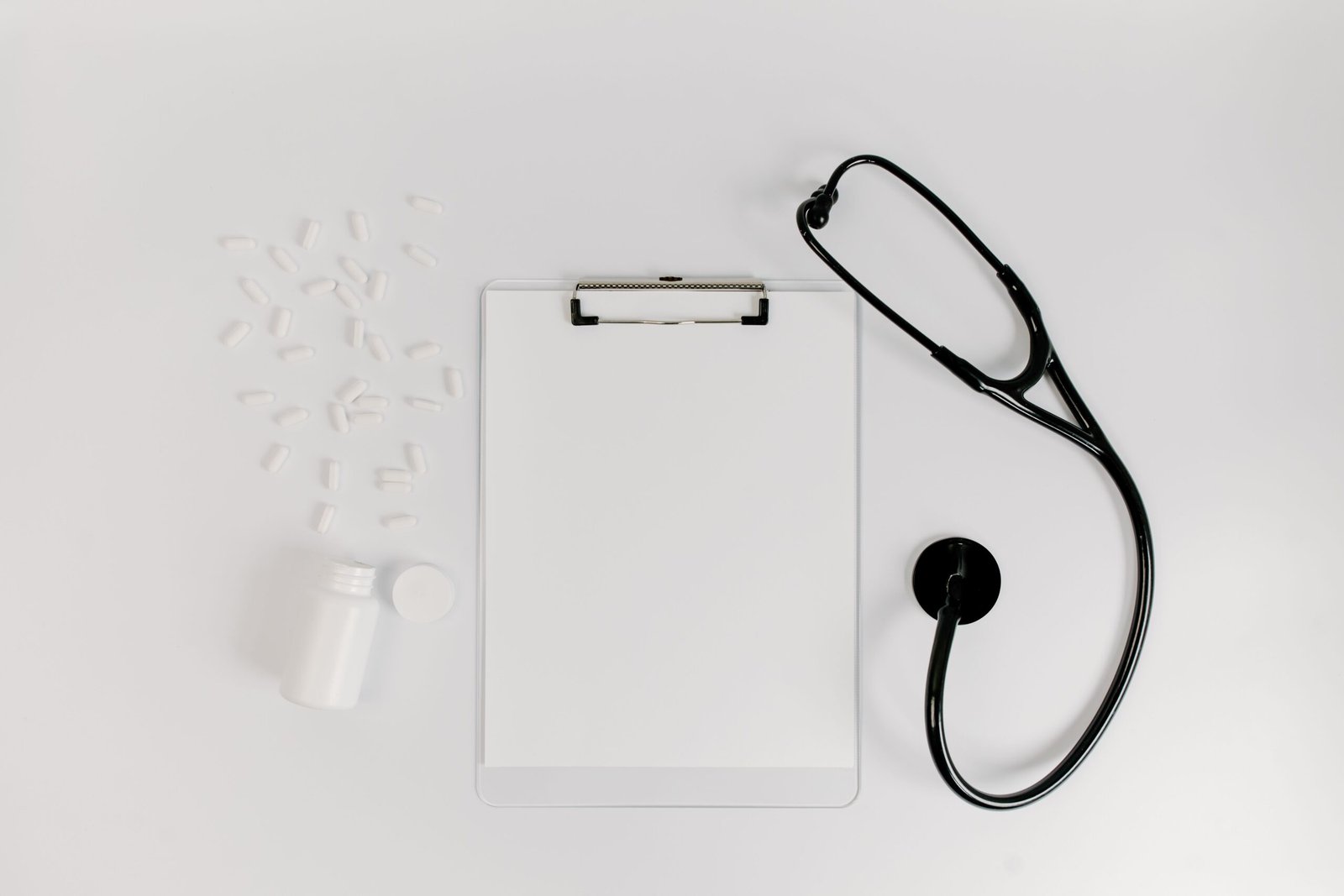Clipboard, prescription bottle, pills, and stethoscope arranged on a white surface.