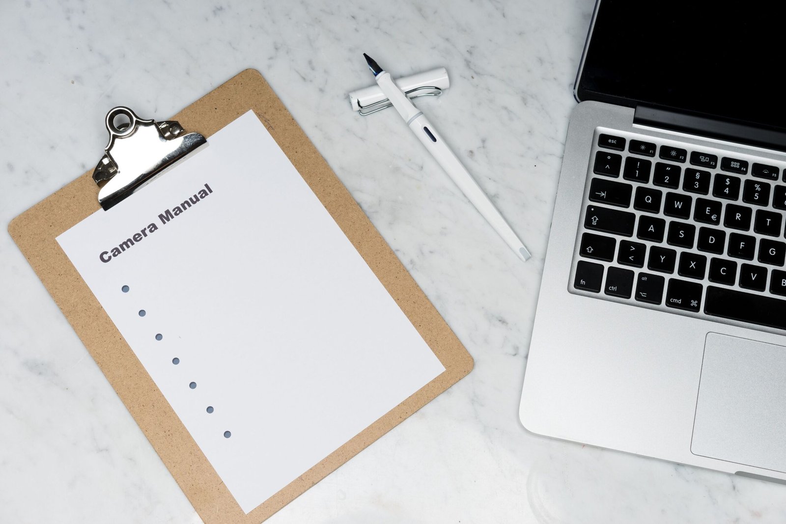 Clipboard, pen, and laptop arranged on a light marble desk.