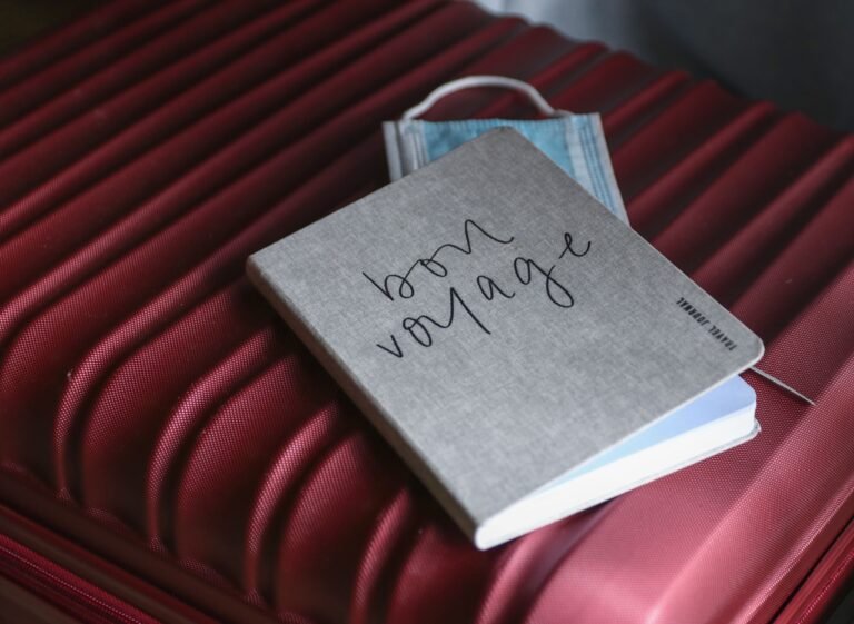 Notebook and face mask resting on a red suitcase prepared for travel.
