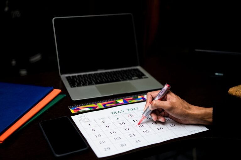 Person marking a date on a calendar at a desk with a laptop and phone.