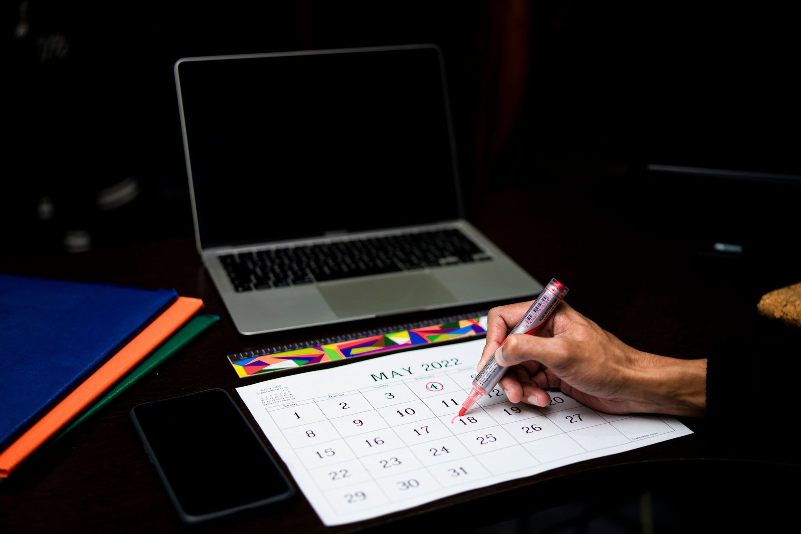 Person marking a date on a calendar at a desk with a laptop and phone.
