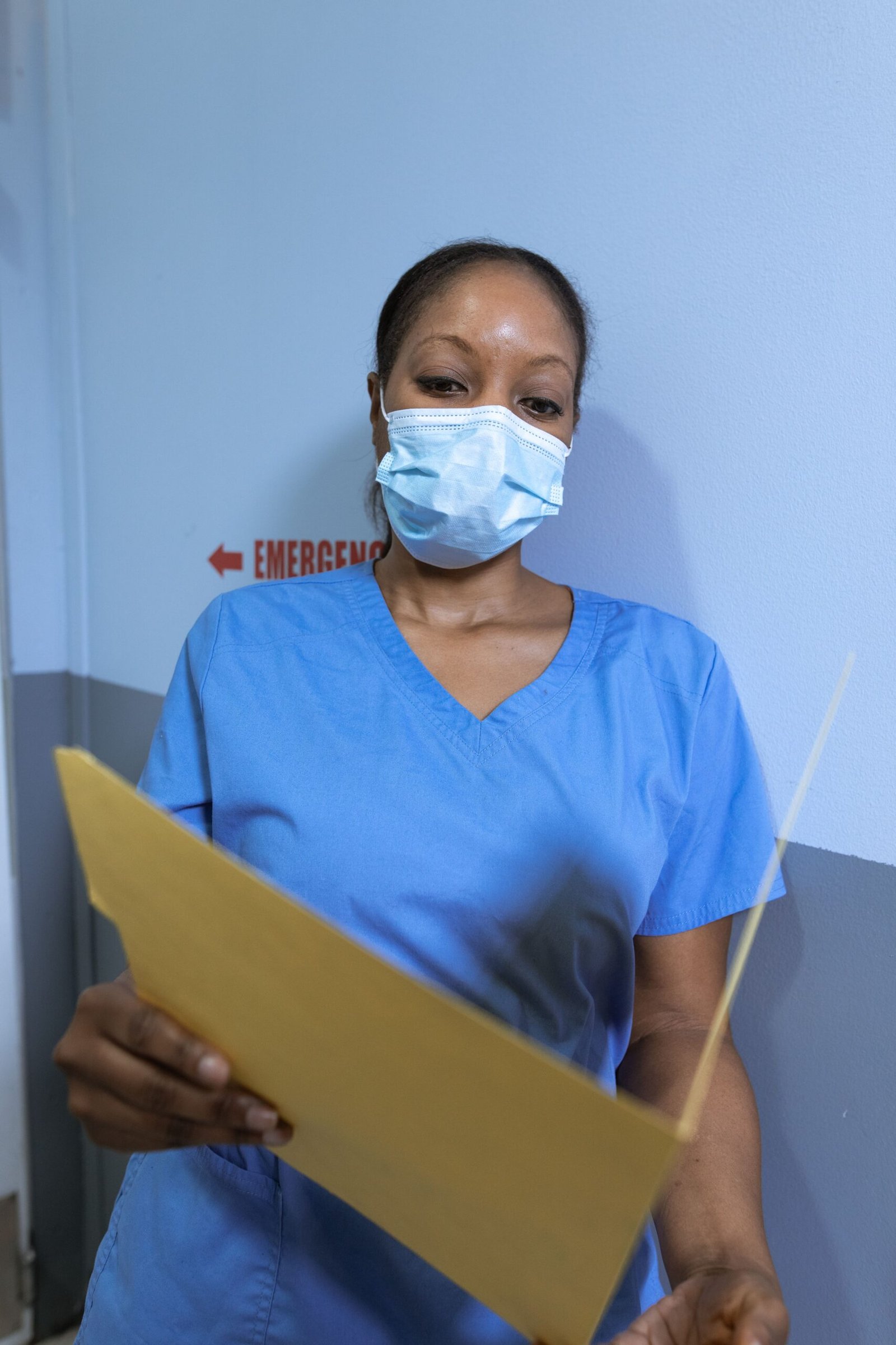 Healthcare professional in blue scrubs and a face mask reading a folder in a corridor.