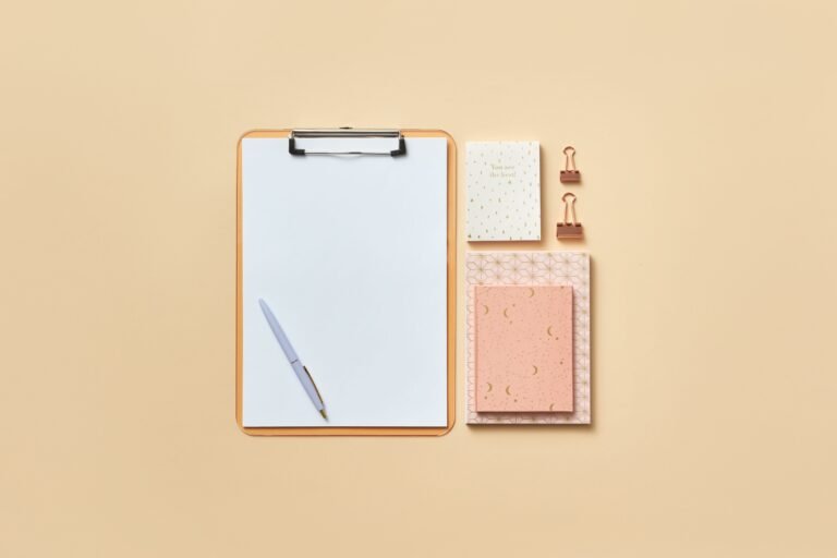 Blank clipboard, pen, notebooks, and paper clips arranged on a beige surface.