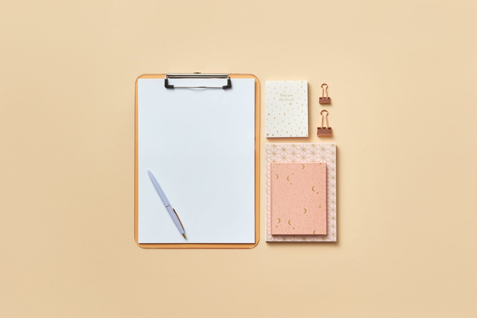 Blank clipboard, pen, notebooks, and paper clips arranged on a beige surface.