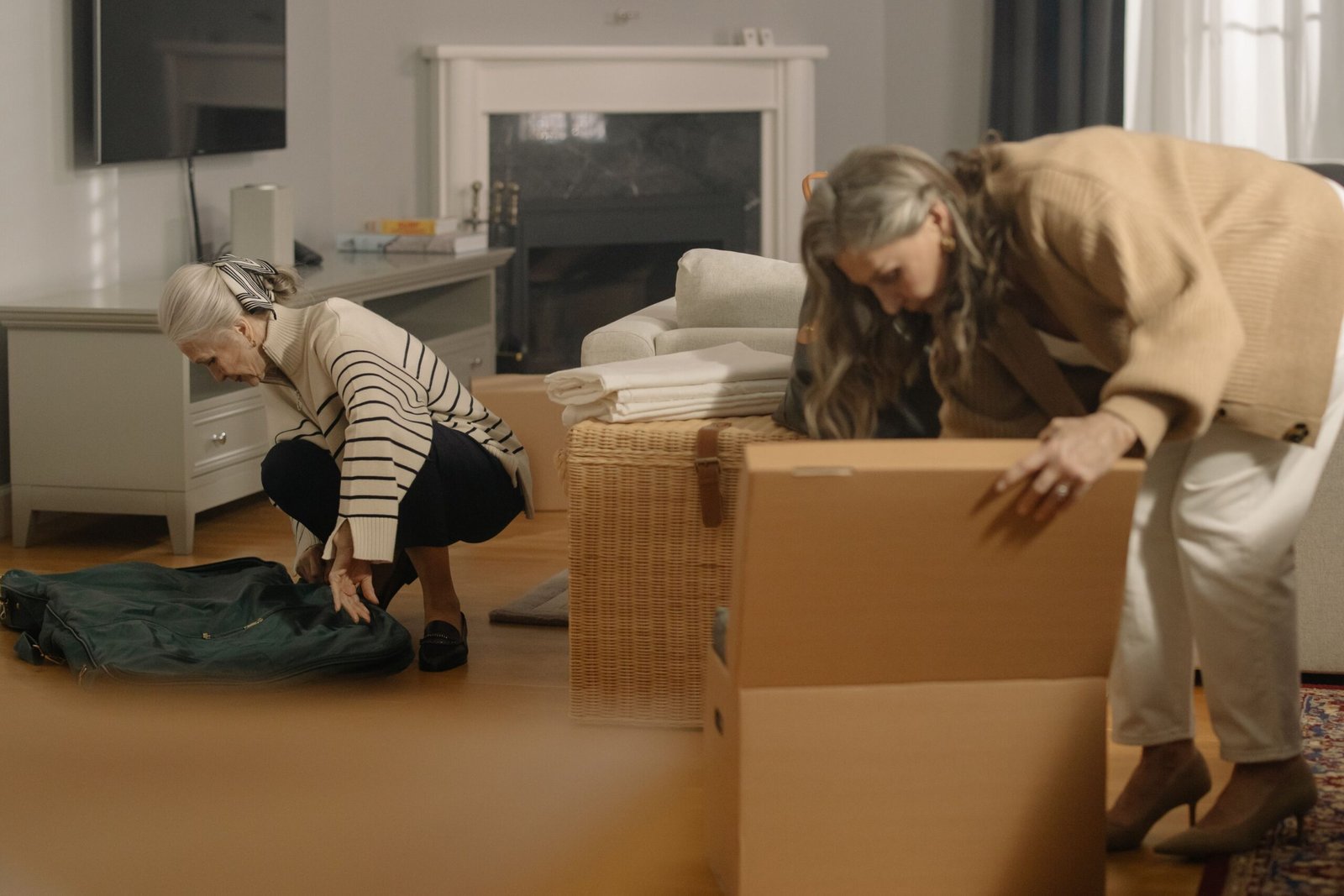 Two older women packing personal belongings into a suitcase and a cardboard box in a living room