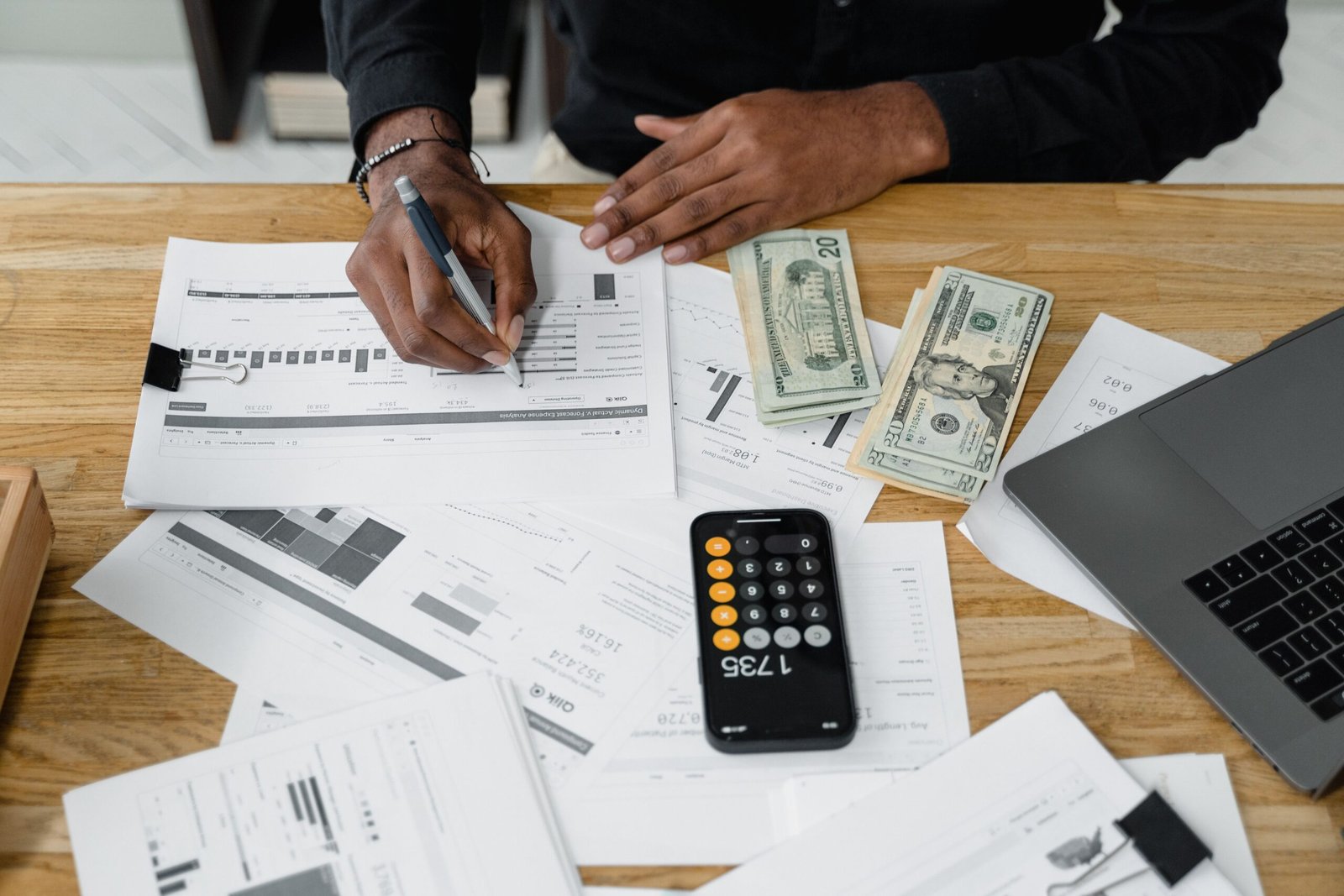 Person reviewing financial reports with a calculator, cash, and laptop.