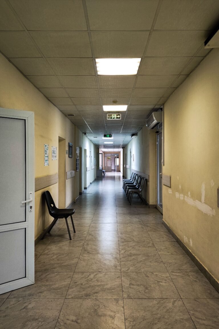 Empty healthcare corridor with open doors and chairs along the wall.