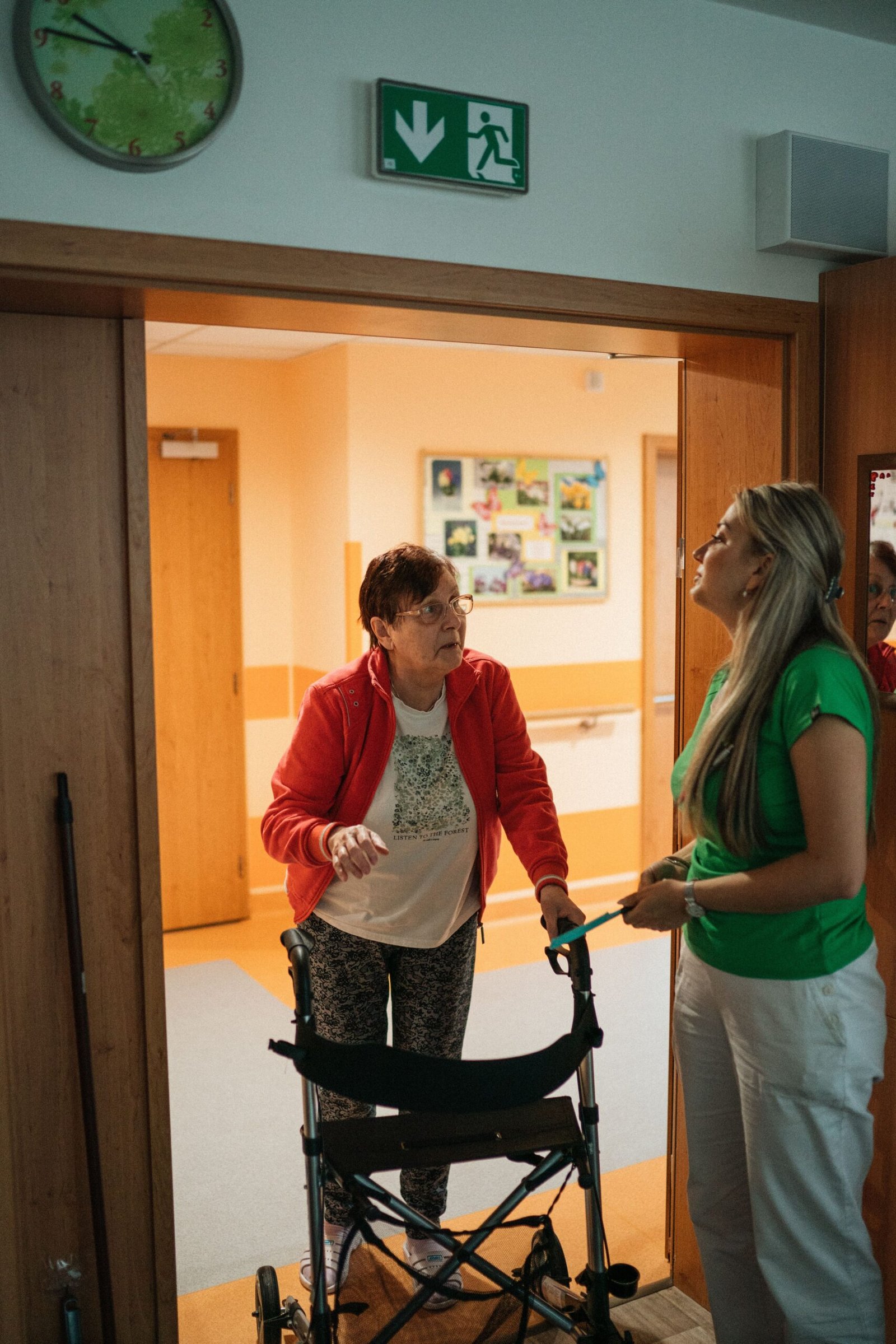 Caregiver standing with an older woman using a walker in a retirement-home doorway.