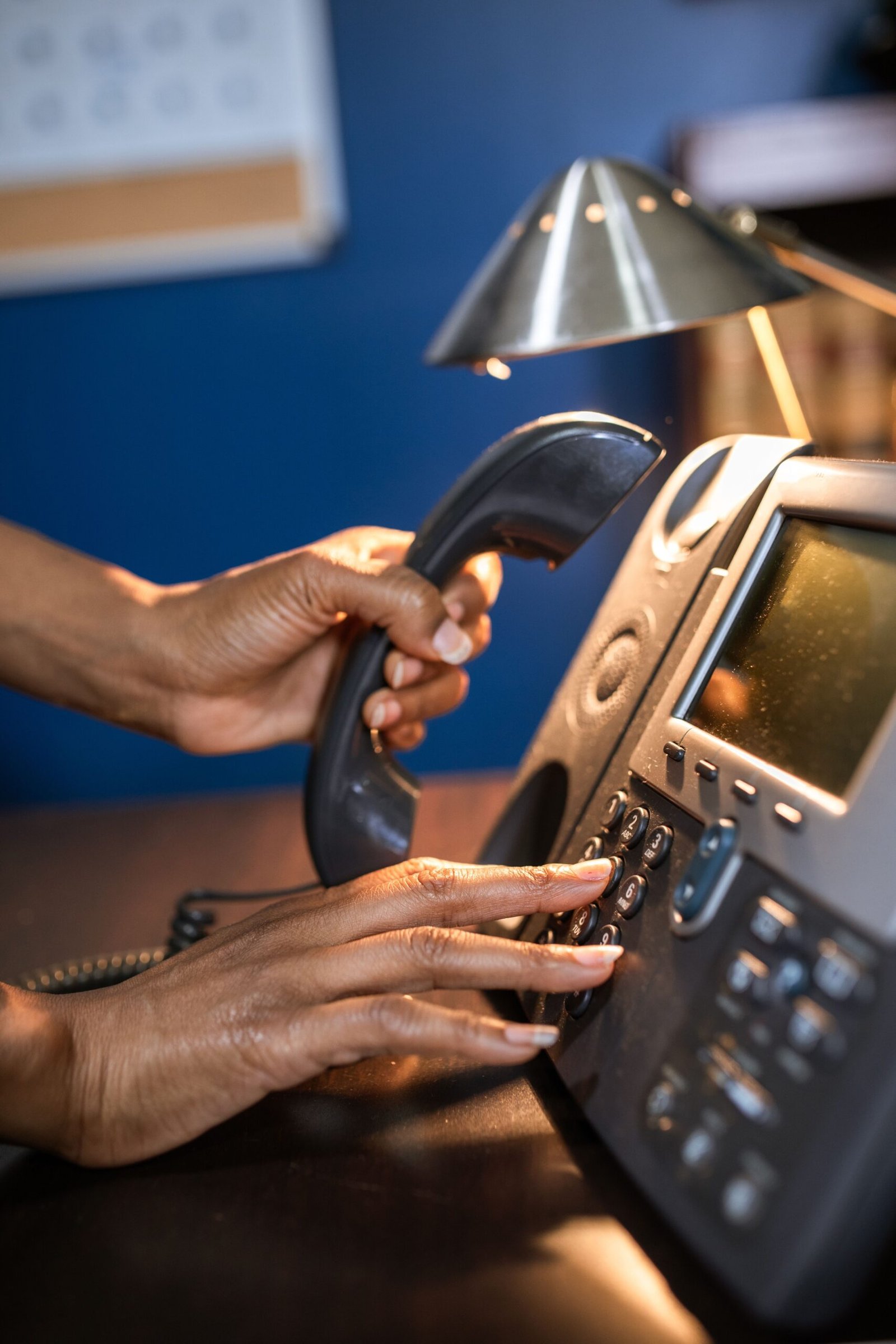 Hands holding a telephone receiver and dialing a desk phone.