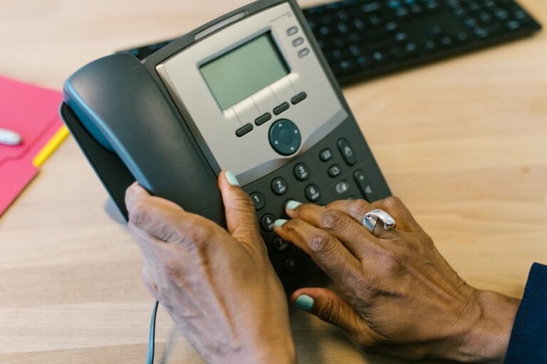 Hands dialing a desk telephone beside a notepad and keyboard.