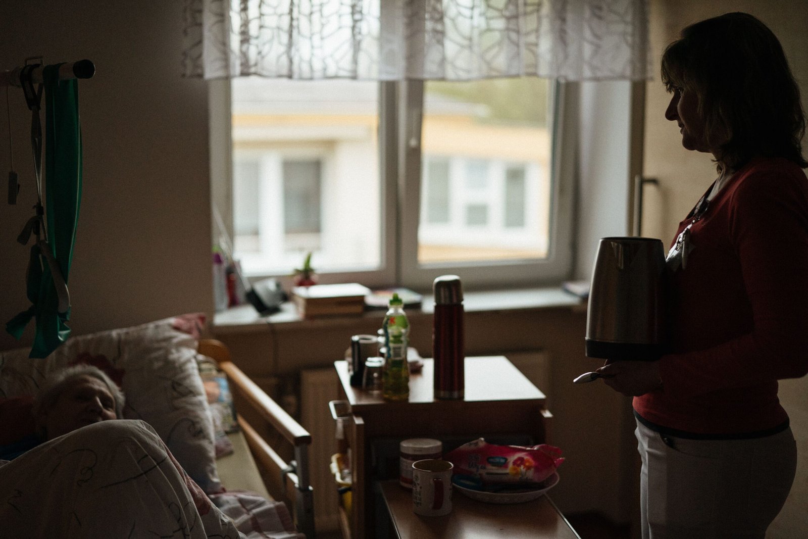 Caregiver supporting an older woman beside her bed in a residential care room