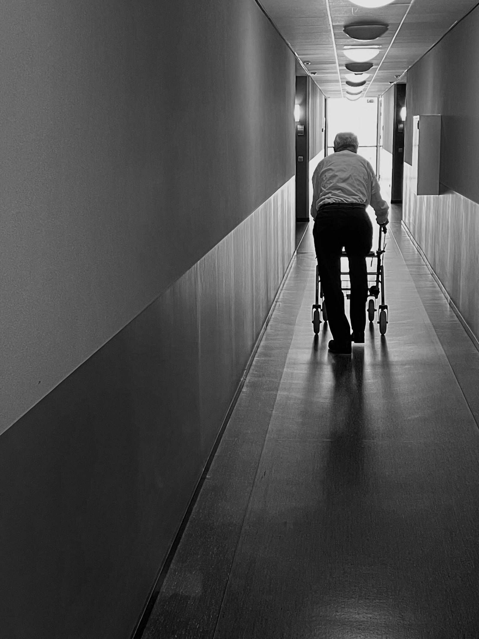 Older adult using a walking aid in a long indoor corridor.
