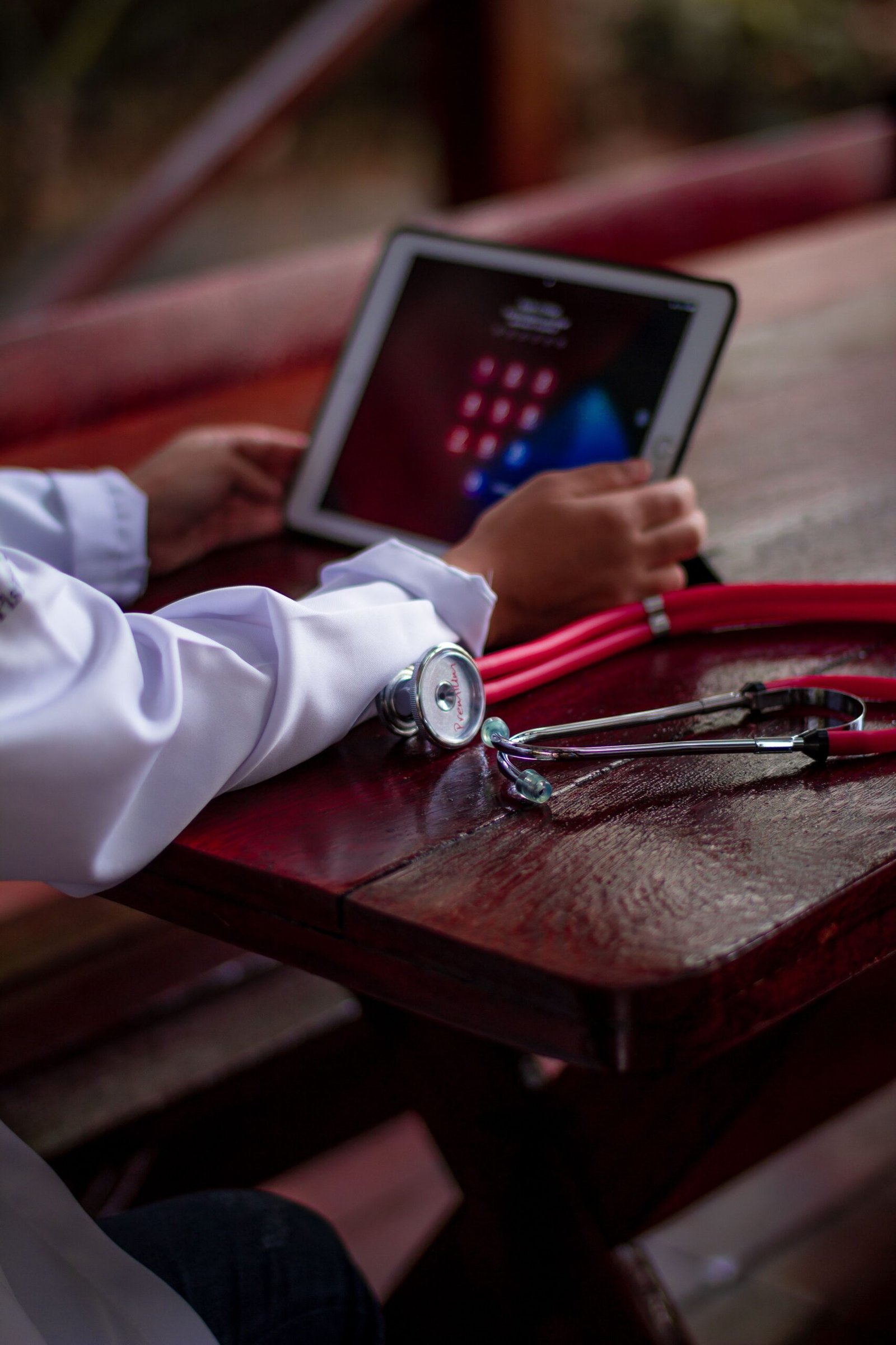 Hands using a tablet beside a red stethoscope on a wooden table.
