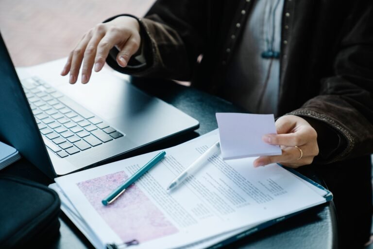 Hands reviewing documents beside an open laptop at a desk.