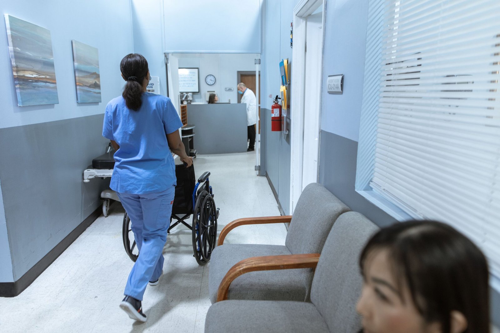 Healthcare worker pushing an empty wheelchair through a bright corridor.