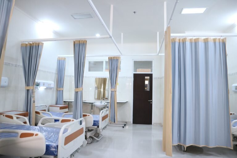 Empty medical beds separated by blue-gray privacy curtains in a bright healthcare room.