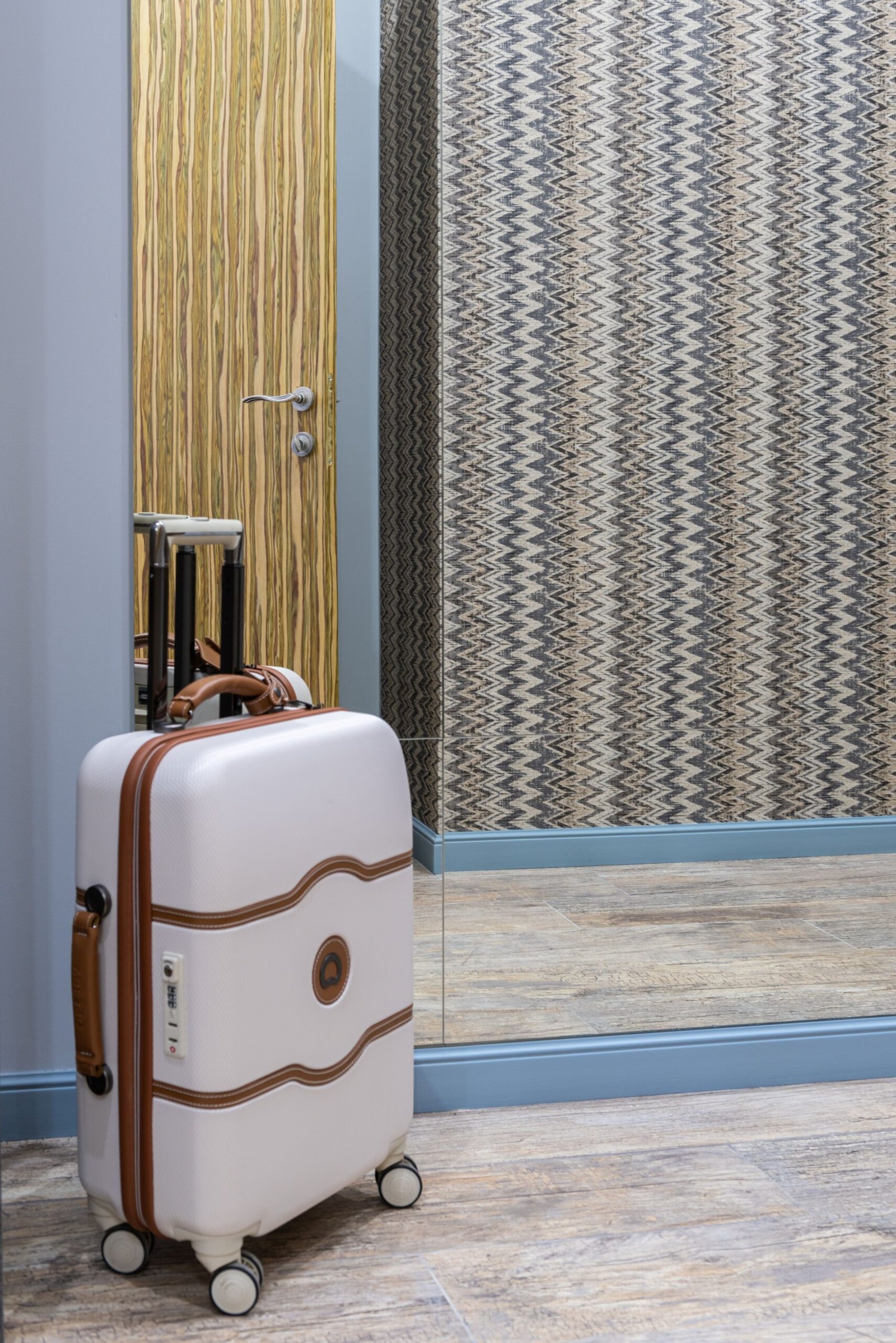 A small wheeled suitcase standing beside a mirror and doorway.