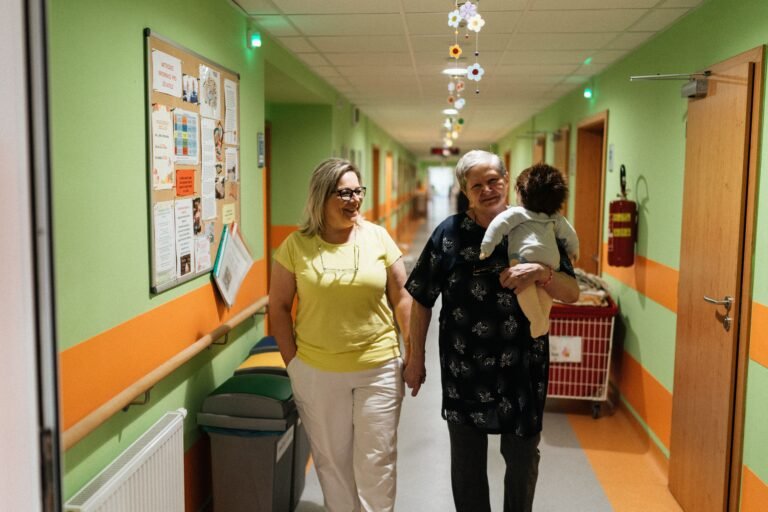 Caregiver walking with an older woman through a nursing-home corridor.